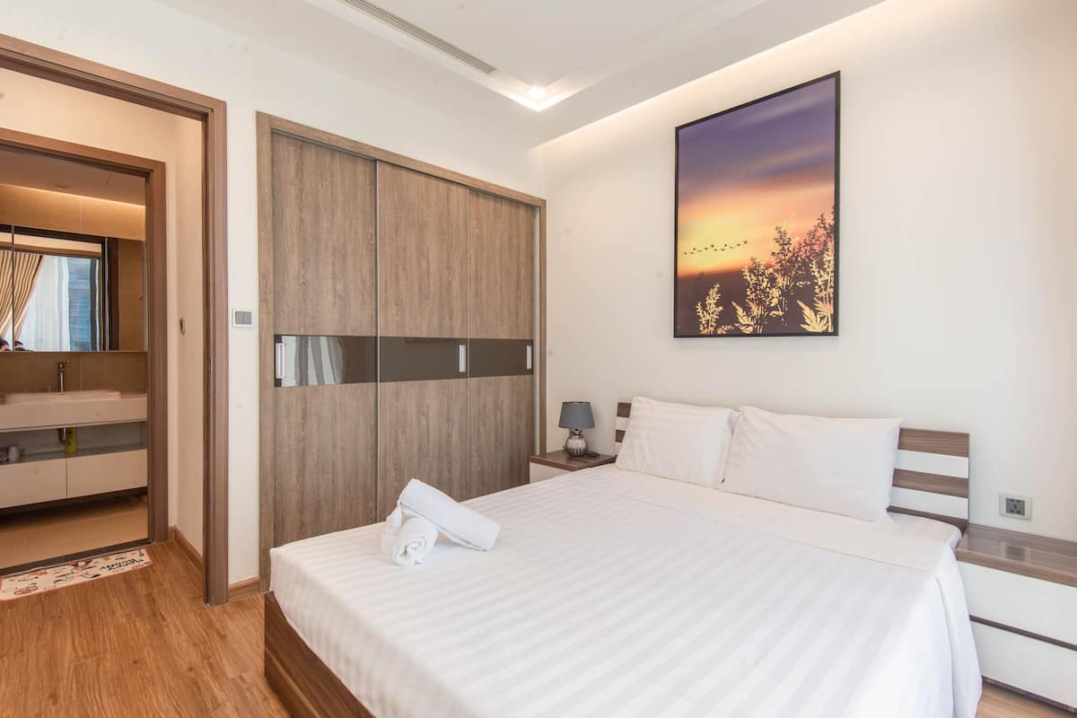 A modern hotel room with a large bed, a wooden wardrobe, a bathroom with a mirror, and a sunset painting on the wall.
