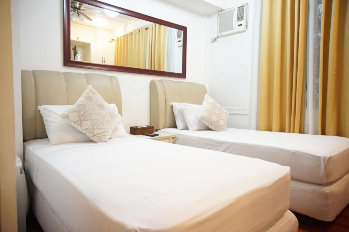 A hotel room with two beds, a bedside table, a mirror, and a window with curtains.