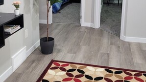 A modern living room with a large plant, a patterned rug, and a blue bed in the background. - Luxury 2 Bedroom Suite (Airdrie)