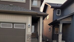 A two-story house with a garage, a front yard with grass, and a green utility box. - Luxury 2 Bedroom Suite (Airdrie)