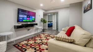 A modern living room with a flat-screen TV mounted on the wall, a beige sofa, a patterned rug, and a small white side table. - Luxury 2 Bedroom Suite (Airdrie)