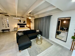 A modern hotel room with a large bed, a sofa, a coffee table, and a dining area visible through a large mirror.