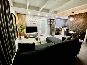 A modern living room with a black sofa, a TV, and a small coffee table. There is a kitchen area with a refrigerator and a dining table.