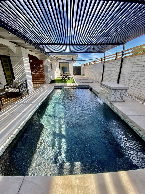 A swimming pool with a modern design, surrounded by a covered patio area with seating and a view of the sky.