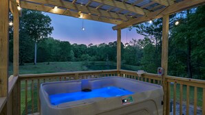 Een hottub onder een houten pergola met lichtjes, omgeven door een houten terras en uitzicht op een bebost gebied.