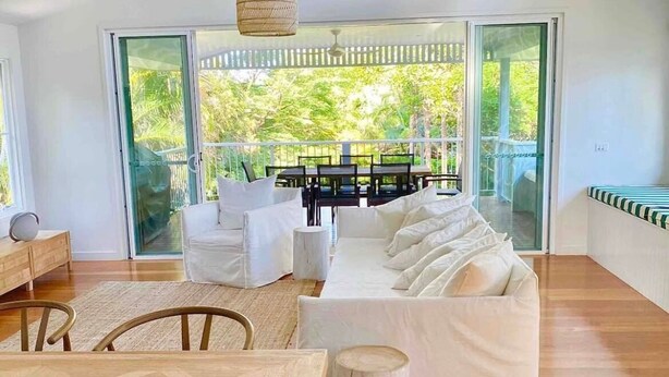 Un salon moderne avec un canapé blanc, une table à manger et des chaises. Une grande porte-fenêtre coulissante donne accès à un balcon offrant une vue sur la verdure.