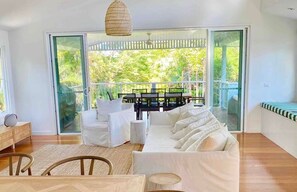 Una sala de estar moderna con un sofá blanco, una mesa de comedor y sillas. Hay un amplio ventanal corredero que da a un balcón con vistas a zonas verdes.