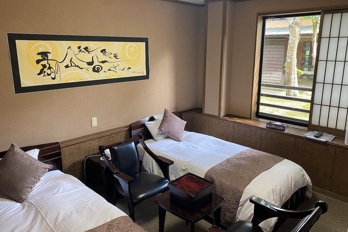 Une chambre d’hôtel avec deux lits, un bureau, une chaise et une grande fenêtre donnant sur l’extérieur.