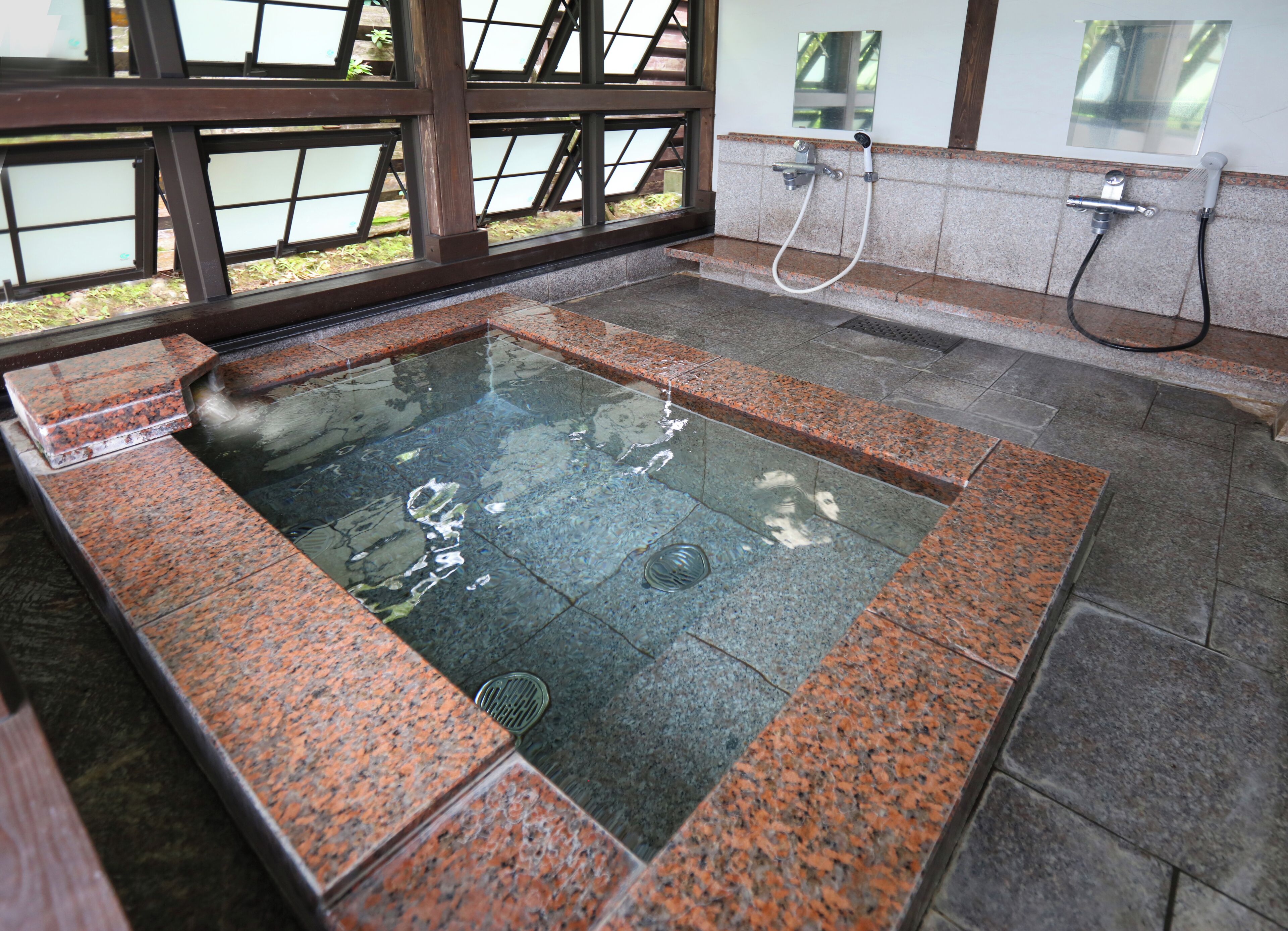 A hot tub with a granite border, a shower area, and a window with a view of greenery.