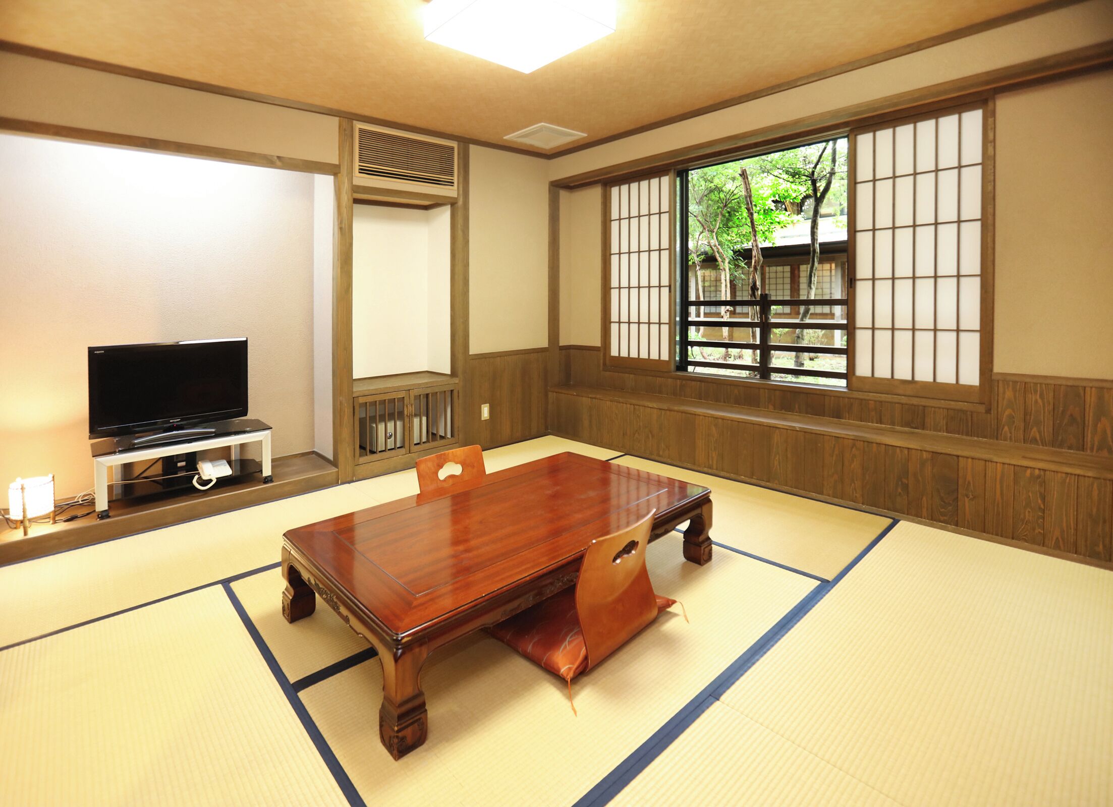 Una habitación japonesa tradicional con suelo de tatami, una mesa de madera baja, un televisor sobre un soporte y una ventana con vistas a la vegetación.