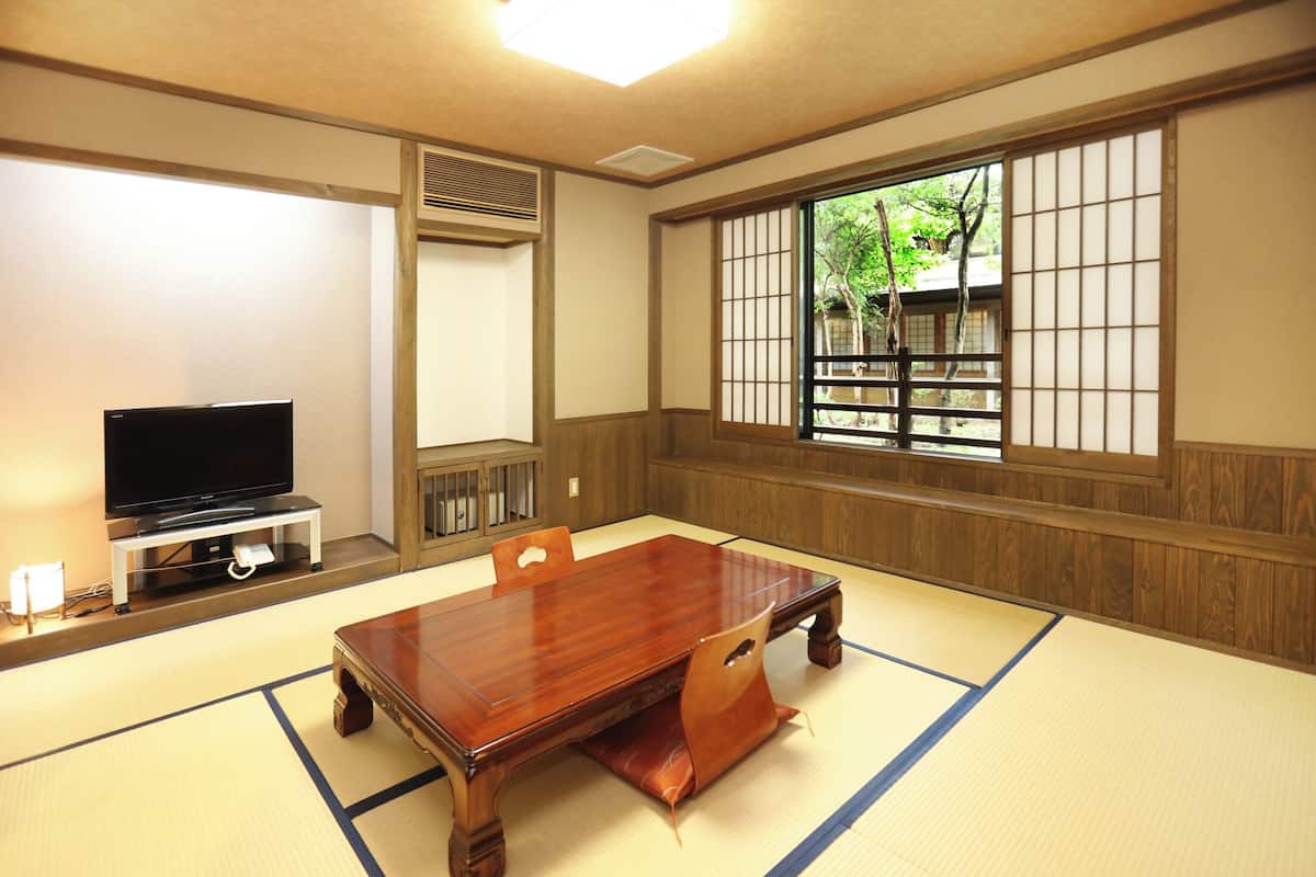 Une pièce de style japonais traditionnel, avec un sol recouvert de tatamis, une basse table en bois, un téléviseur posé sur un meuble et une fenêtre donnant sur un paysage verdoyant.