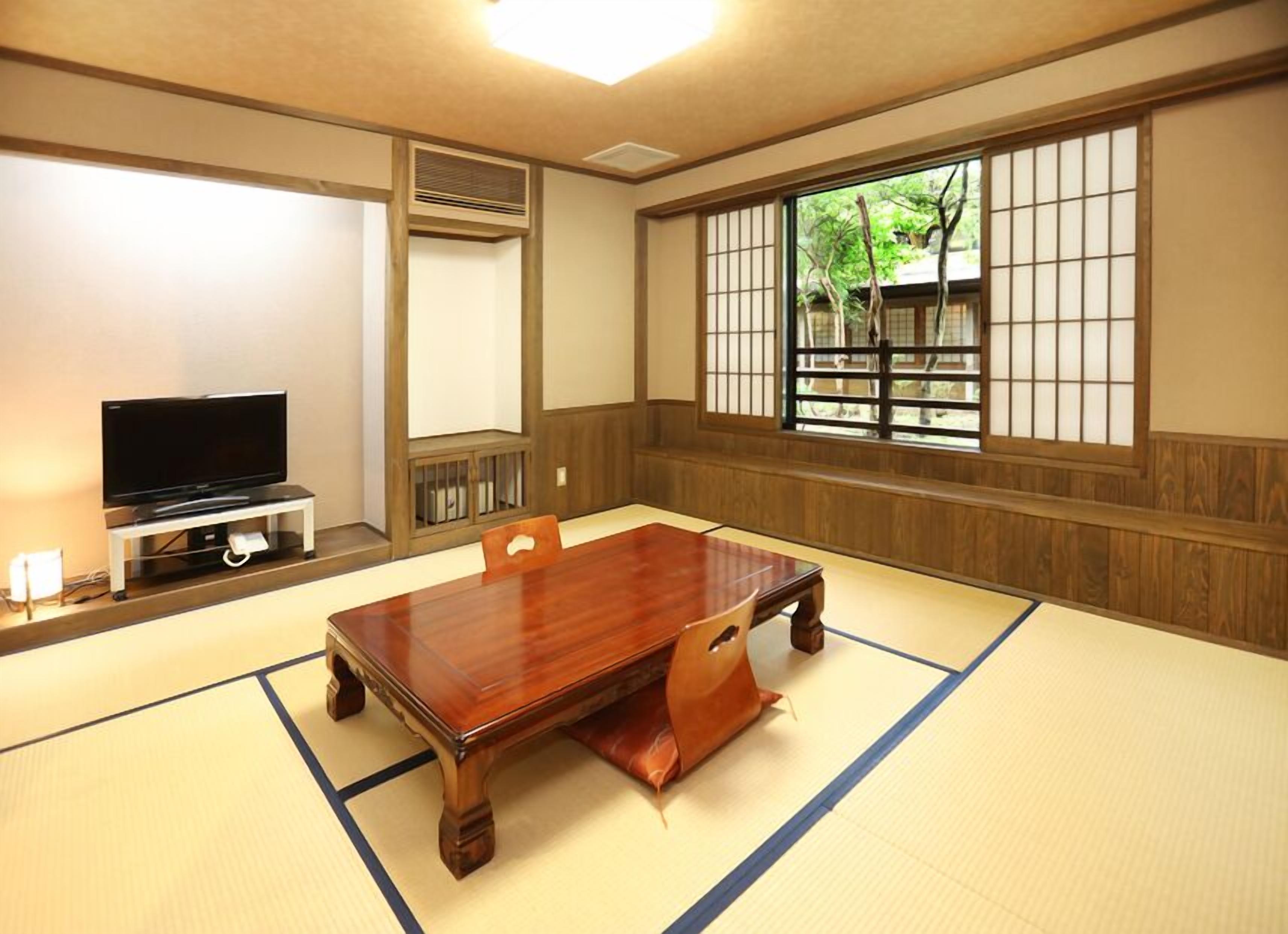 Een traditionele kamer met tatami matten, een houten tafel, een televisie op een standaard en een raam met uitzicht op groen.