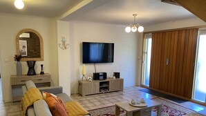 A modern living room with a grey sofa set, a wooden cabinet with a flat-screen TV, and a large sliding door. - Dromana Charmer - Large family home, 8 min easy walk to the beach. (Dromana)
