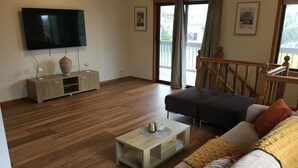 A modern living room with a flat-screen TV, wooden flooring, a sofa set, a coffee table, and a staircase leading to a balcony. - Dromana Charmer - Large family home, 8 min easy walk to the beach. (Dromana)