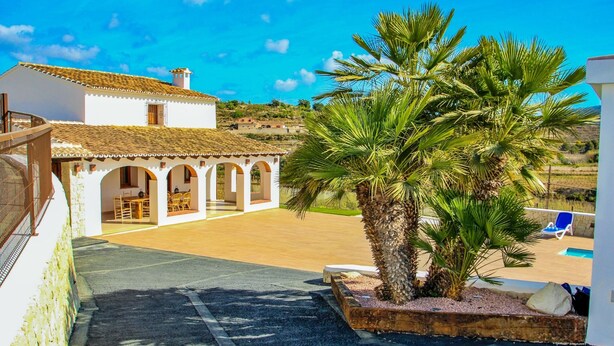 A white building with a tiled roof, a covered patio with chairs, and a palm tree. - Rosalia - holiday home with panoramic view and private pool in Teluda (Teulada)