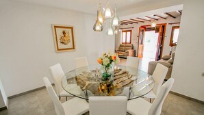 A modern dining area with a glass table surrounded by white chairs, a vase of flowers, and pendant lights. - Rosalia - holiday home with panoramic view and private pool in Teluda (Teulada)