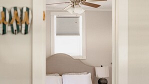 A bedroom with a bed, a ceiling fan, a window with blinds, and a nightstand with a lamp.