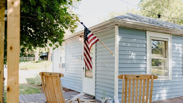 A small wooden house with a porch, two wooden chairs, and an American flag.