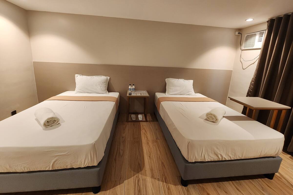 A hotel room with two single beds, a small bedside table with toiletries, a wooden floor, and a window with curtains.