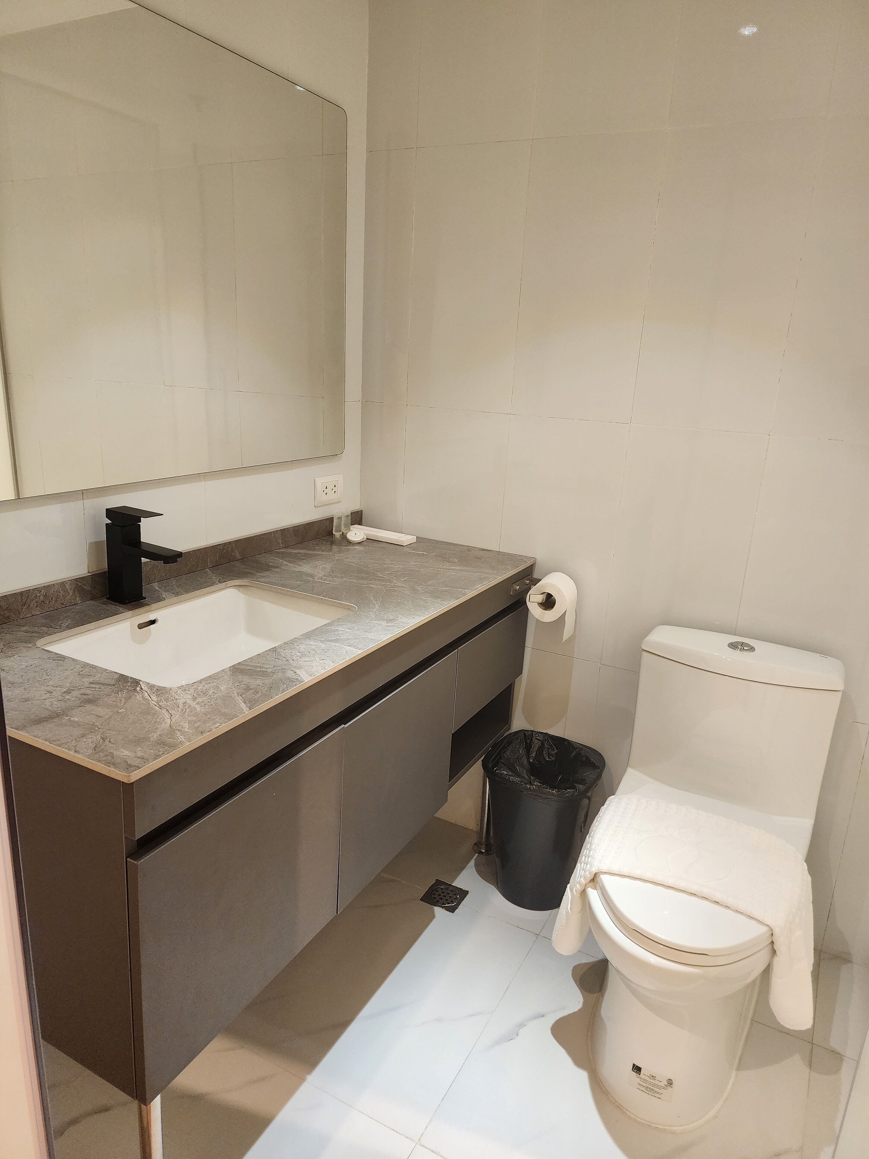 A bathroom with a sink, mirror, toilet, and trash bin.