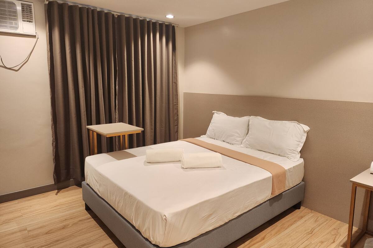 A hotel room with a bed, a small table, a curtain, and an air conditioning unit.