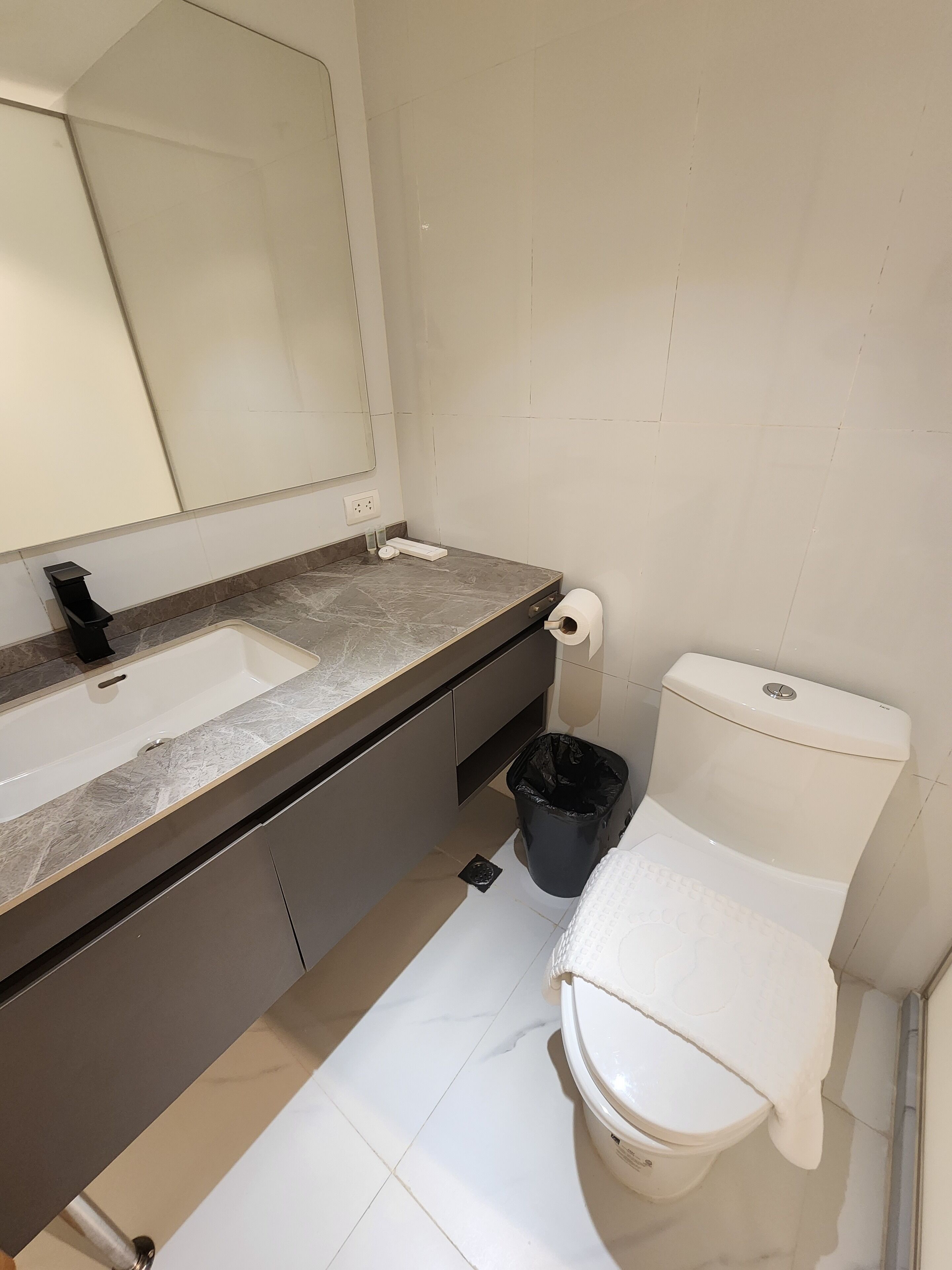 A bathroom with a sink, mirror, toilet, and trash bin.