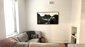 Uma sala de estar moderna com sofá seccional, um banquinho pequeno e uma pintura emoldurada de uma paisagem montanhosa na parede.