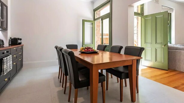 Uma área de refeições com uma mesa de madeira e cadeiras pretas, uma cesta de frutas sobre a mesa e uma porta verde ao fundo.