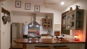 A kitchen with a central island, wooden stools, a stove, a refrigerator, and a cabinet with glass doors. - Romantic rainforest retreat with magical waterfall (Cow Bay)