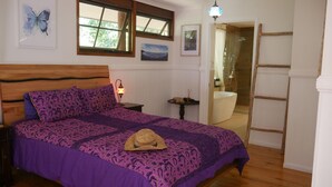 A bedroom with a wooden bed, purple bedding, a nightstand with a lamp, a window with a view, and a bathroom visible through an open door. - Romantic rainforest retreat with magical waterfall (Cow Bay)