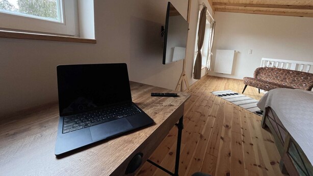 A room with a wooden desk, a laptop, a bed with a patterned cover, a radiator, and a window with curtains. - Kutscherhaus / Stadthaus Usedom (Usedom)