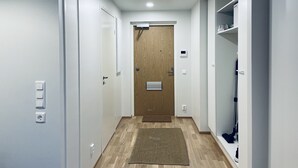 A hallway with a wooden door, white walls, and a carpet. There are white cabinets and a shelf with items on the right side. - Completely new designer apartment on the ground floor with a spacious patio (Espoo)