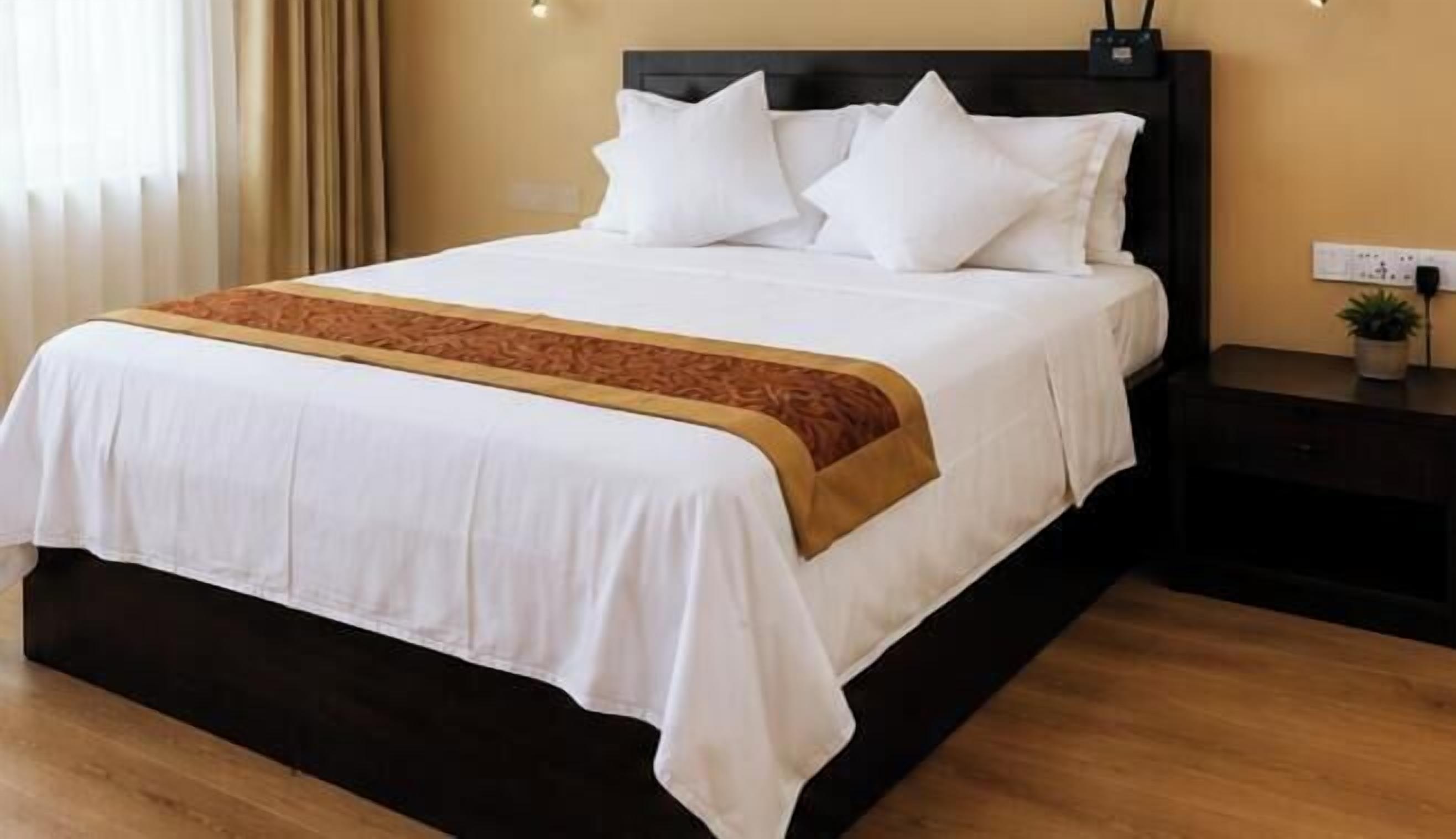 A neatly made bed with white bedding and a brown accent, a bedside table with a lamp, and a power outlet on the wall.
