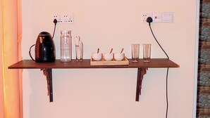 A kitchen area with a wooden shelf holding a kettle, glasses, and bowls. There is a power outlet on the wall and a small appliance below the shelf. - Woody Crest Hotel (Trincomalee)