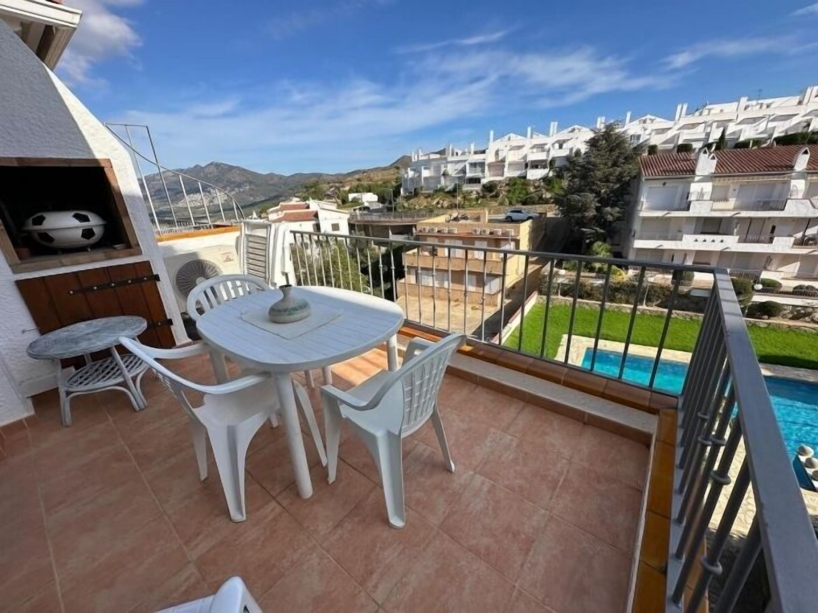 Een balkon met een tafel, stoelen, een barbecue en uitzicht op een woonwijk met huizen en een zwembad.