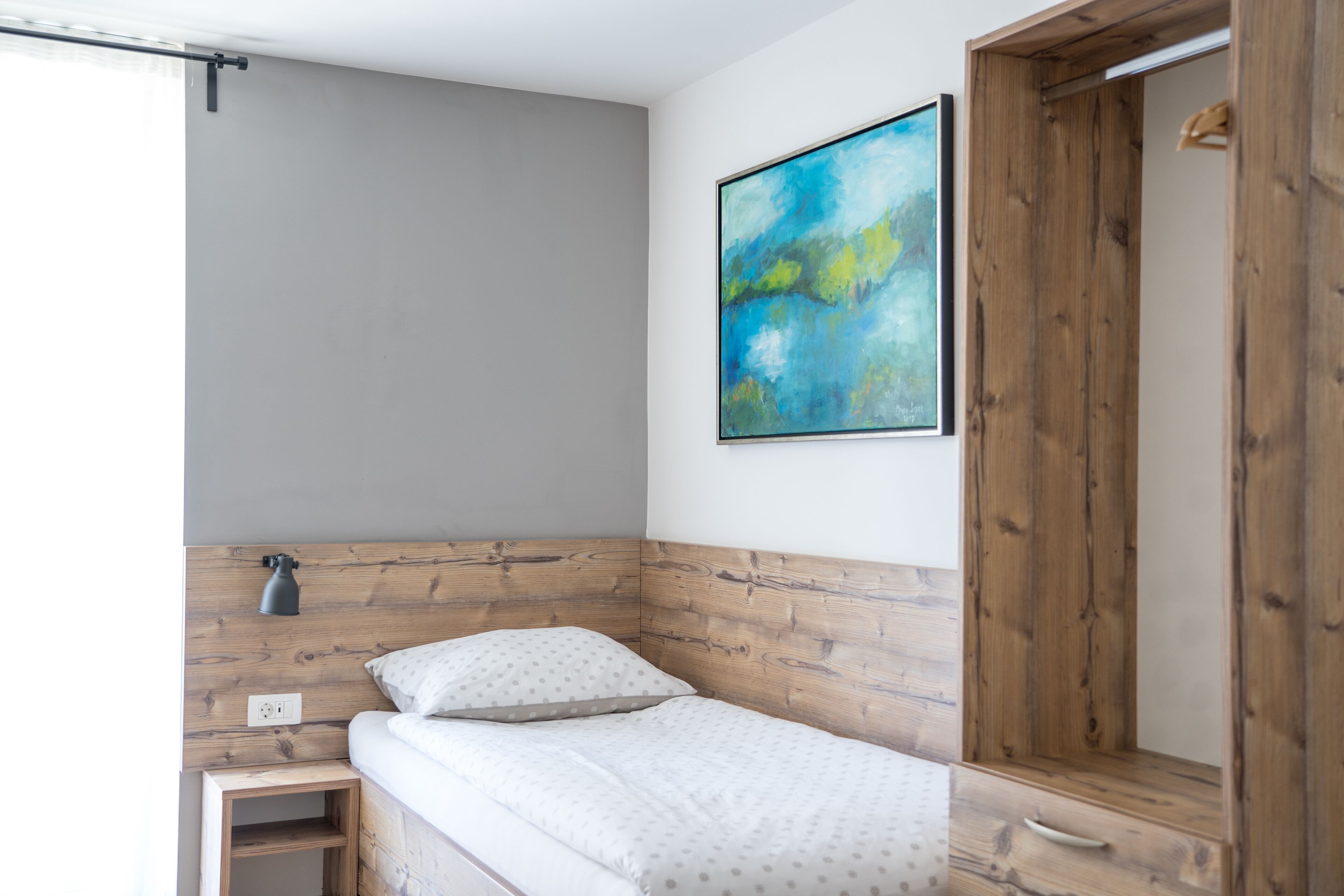A small, neatly arranged bedroom with a single bed, a wooden headboard, a wall-mounted light, and a large abstract painting.