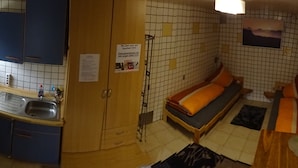 Ett litet rum med ett pentry, en säng med en orange kudde, en liten matta och en tavla som hänger på väggen.