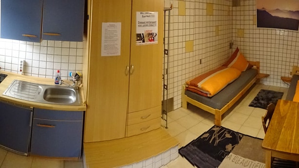 Ett litet, kompakt rum med ett pentry, en säng med en orangef ärgad kudde och ett litet bord.