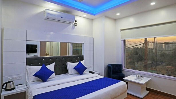 A modern hotel room with a large bed, a bedside table, a nightstand, a window with a city view, and an air conditioning unit. - Hotel Fortune Stay (Ghaziabad)