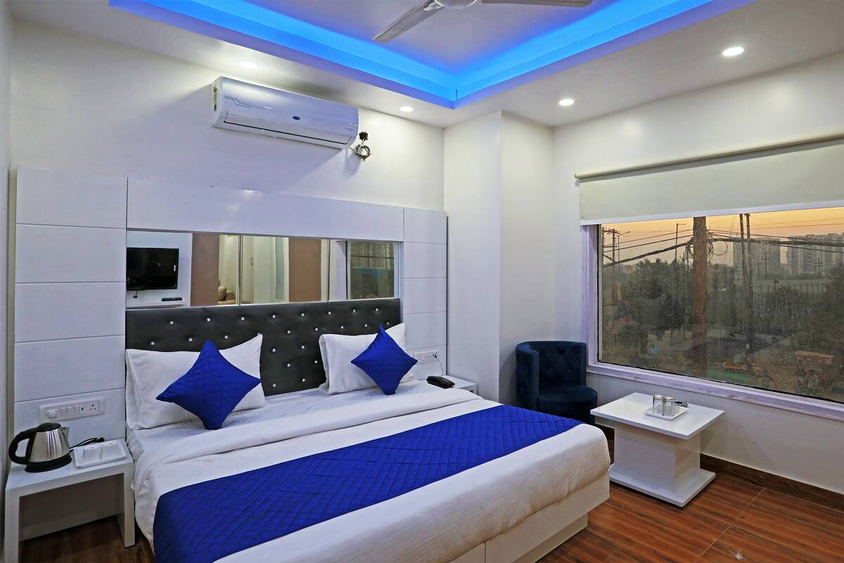 A modern hotel room with a large bed, a bedside table, a nightstand, a window with a city view, and an air conditioning unit.