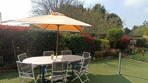 Um pátio externo com mesa e cadeiras redondas, um guarda-sol, e um jardim com plantas variadas e gramado.
