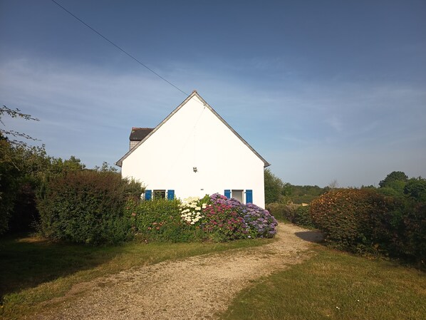 A white building with blue shutters, surrounded by lush greenery and a gravel path. - Country house in Nevez, near Port-Manec'h (Névez)