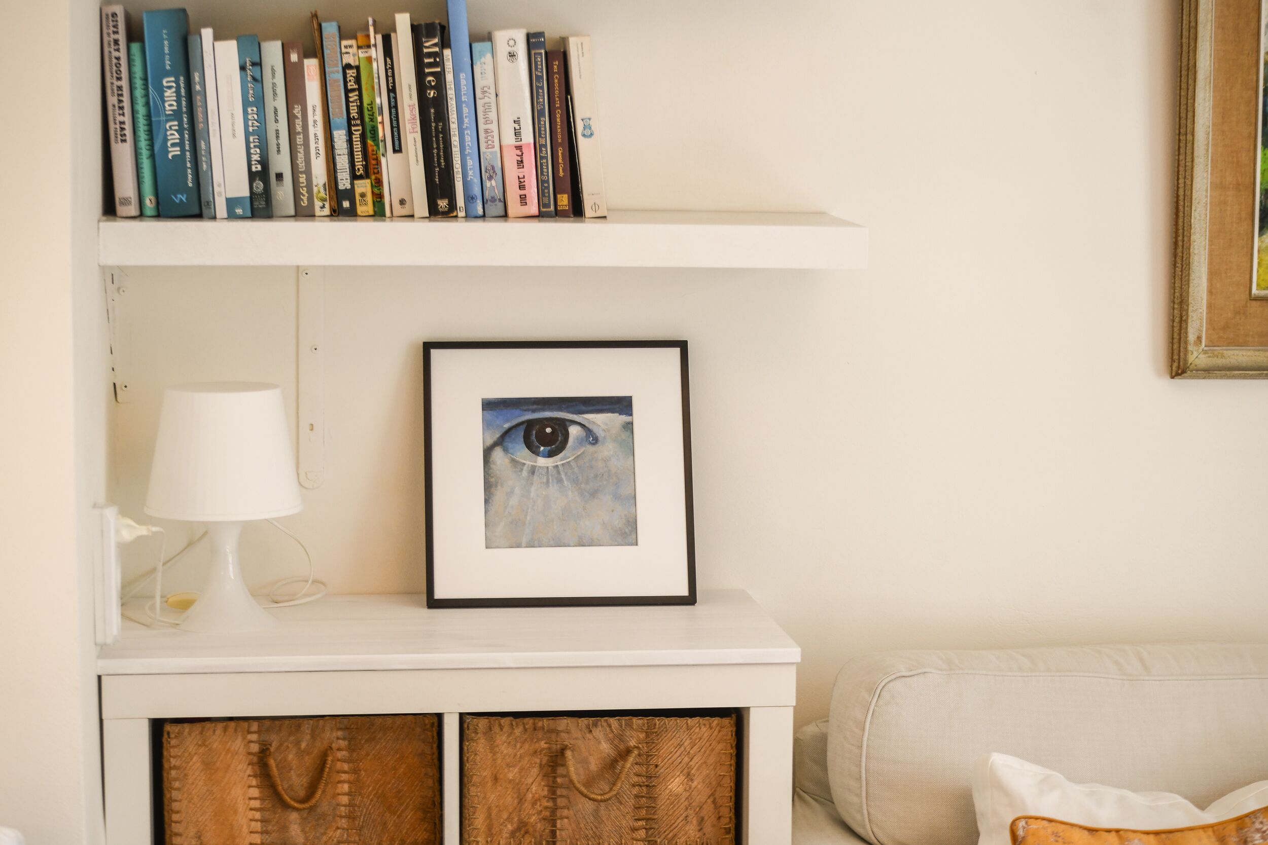 Una estantería con libros, un cuadro con la imagen de un ojo, una lámpara blanca y un aparador de madera.
