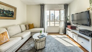 Un salon moderne avec un canapé d’angle, une table basse ronde, un téléviseur à écran plat et une grande fenêtre agrémentée de rideaux.