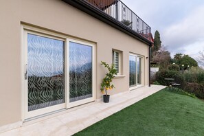 Uma casa moderna com amplas portas de vidro deslizantes, um pequeno jardim com gramado e um vaso de planta, e uma varanda com guarda-corpo.