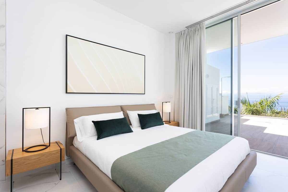 Una habitación de hotel moderna con una cama grande, dos mesitas de noche, un amplio ventanal con cortinas y vistas al mar.