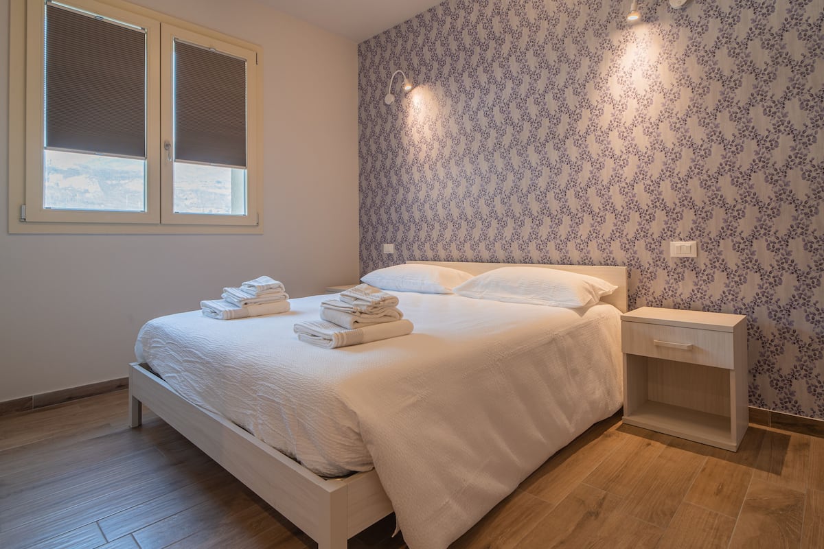 A bedroom with a bed, a nightstand, a window with blinds, and a wall with a floral pattern.