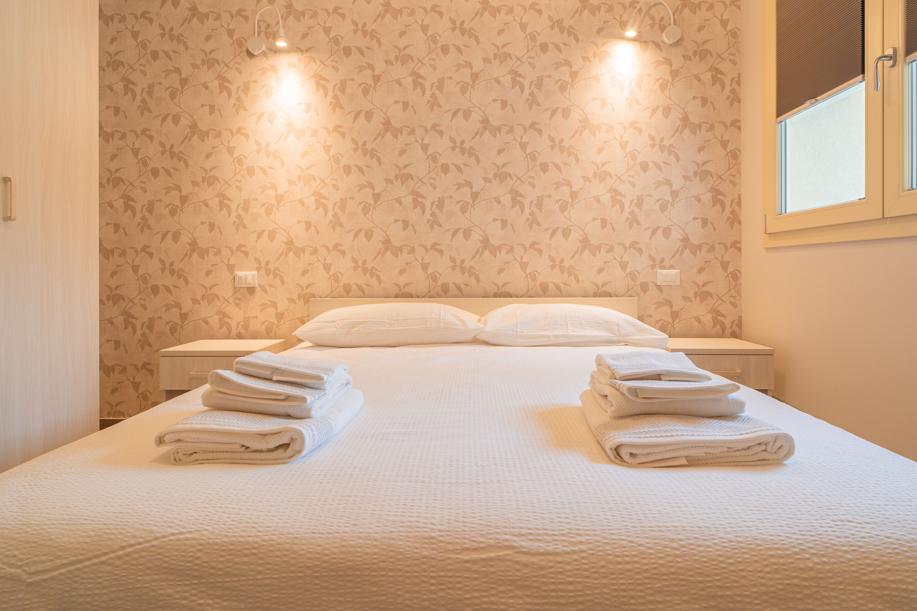 Ein ordentlich bezogenes Bett mit gefalteten Handtüchern, ein Nachttisch und eine Wand mit floralem Muster.
