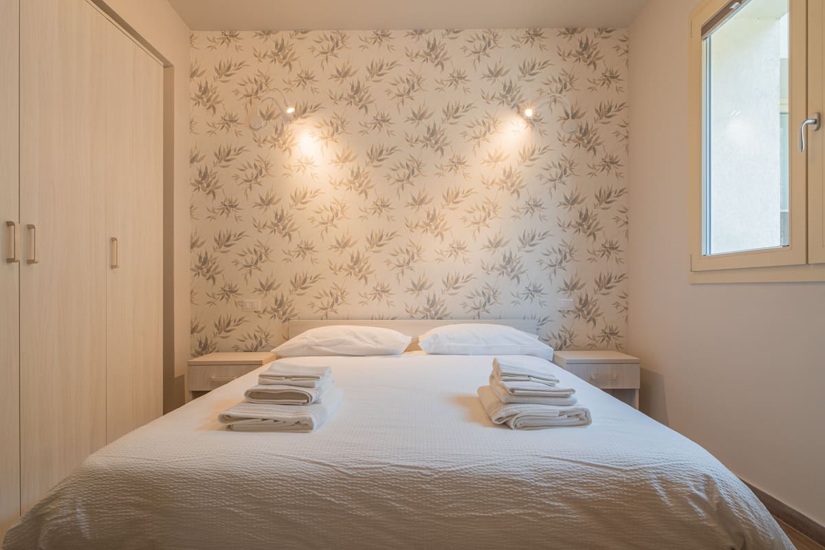 A neatly made bed with folded towels, a wooden wardrobe, and a floral wallpaper pattern.