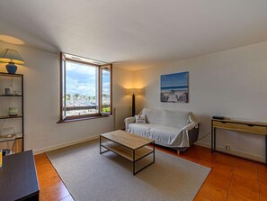 Un salon moderne comprenant un canapé, une table basse, une lampe, un bureau et une vue sur le port de plaisance, visible à travers une grande fenêtre.